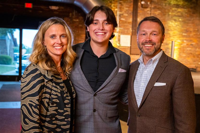 A young man wearing a grey checked blazer smiles with a woman wearing a patterned top and a man wearing a brown blazer